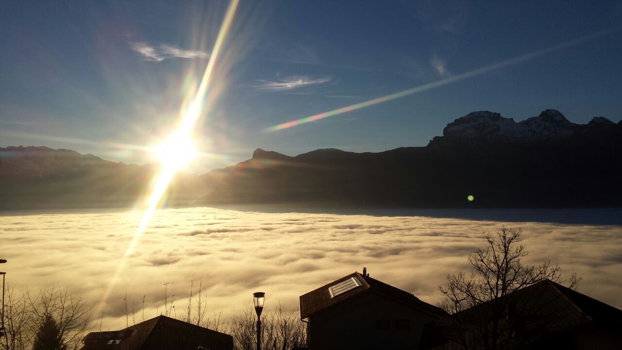 Die Sonne steht über Bergen und beleuchtet ein Meer aus Nebel mit Hausdächern im Vordergrund.