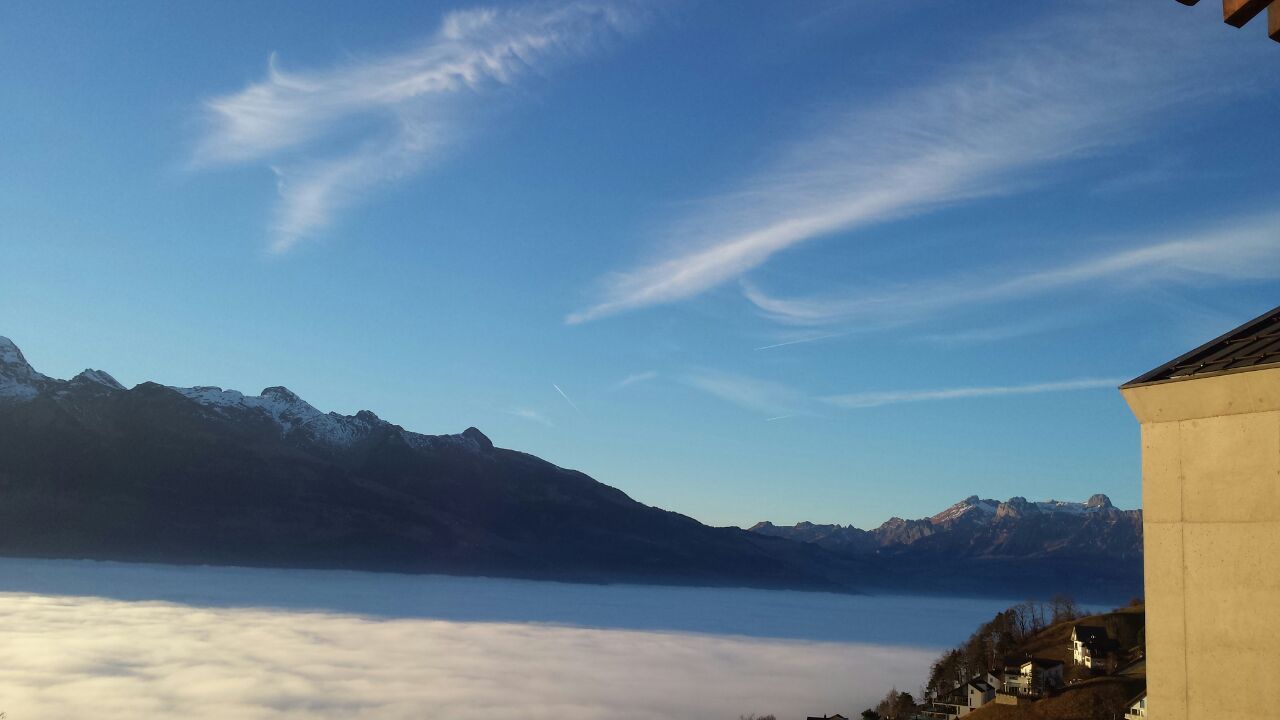 Berge ragen über einer dichten Nebelschicht unter blauem Himmel mit zarten Wolken auf.
