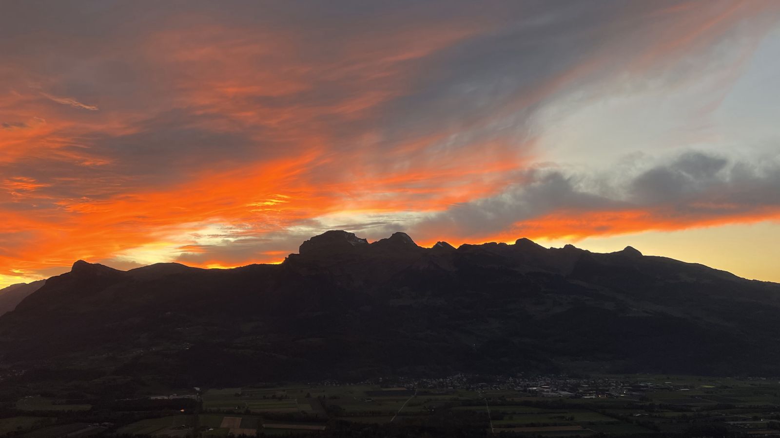 Leuchtender Sonnenuntergang mit orangefarbenen Wolken über einer dunklen Bergsilhouette und einem Tal.