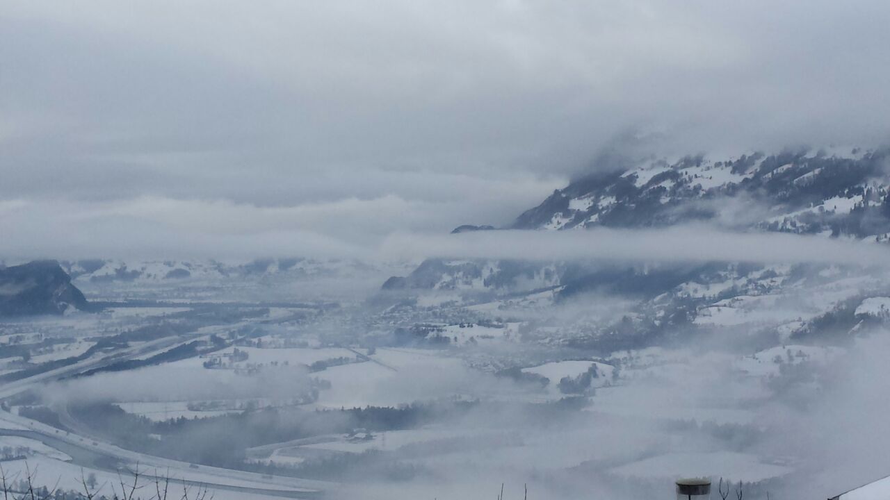 Verschneite Hänge und ein Tal liegen unter tief hängenden Wolken und Nebel.